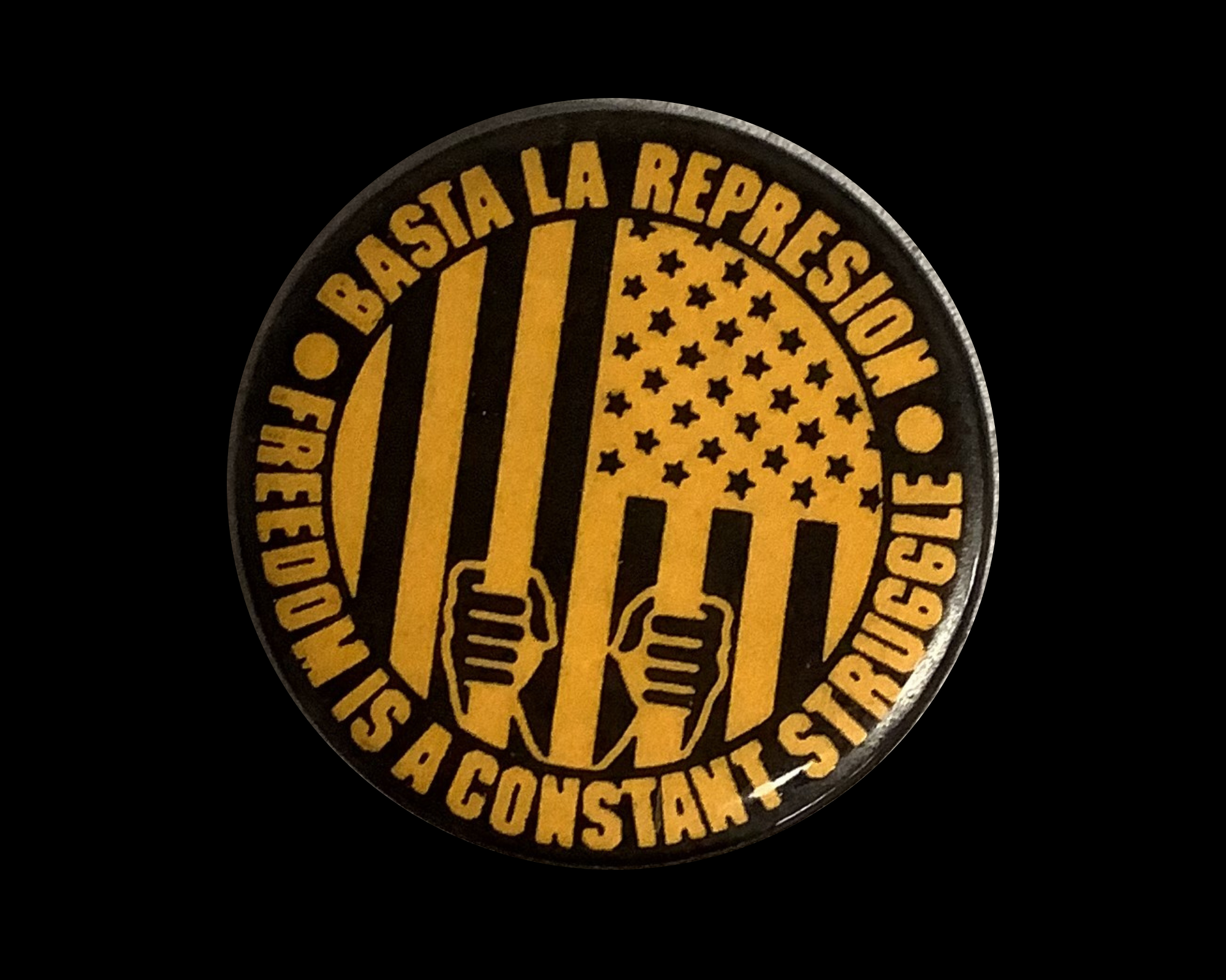 Issued by the National Alliance Against Racist and Political Repression, depicts the logo for the Chicago Alliance Against Racist and Political Oppression. The design in yellow and black depicts the American flag turned vertically, representing prison bars. Hands grip the bars near the botton of the design. The text in yellow reads Basta La Represion [Enough Represion) Freedom is a Constant Struggle.
1 3/4 inch round, metal button with pin back.