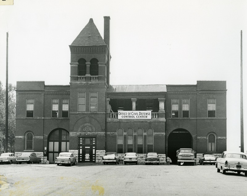 Historic photo of brick building with Office of Civil Defense sign and cars parked nearby.
