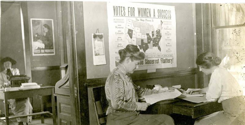Photograph of Women Seated at Desks, with a "Votes For Women A Success" Poster
